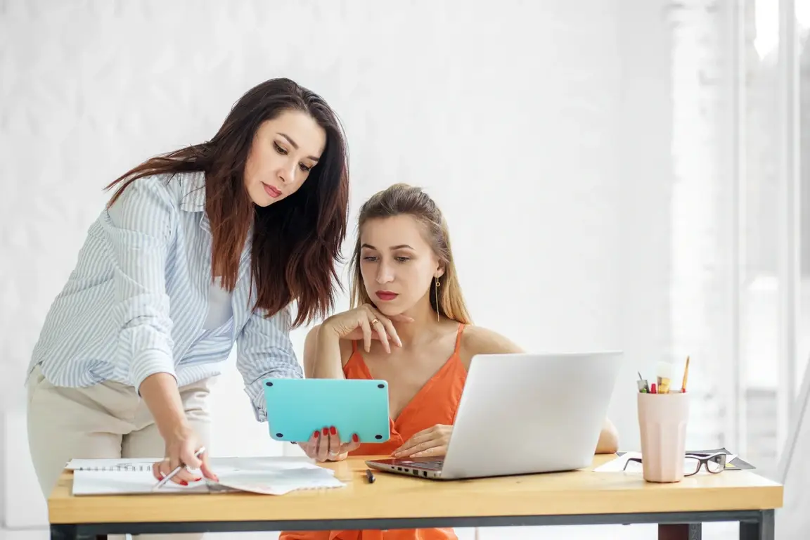 Two women at a desk looking over computer and tablet screens as well as documents.