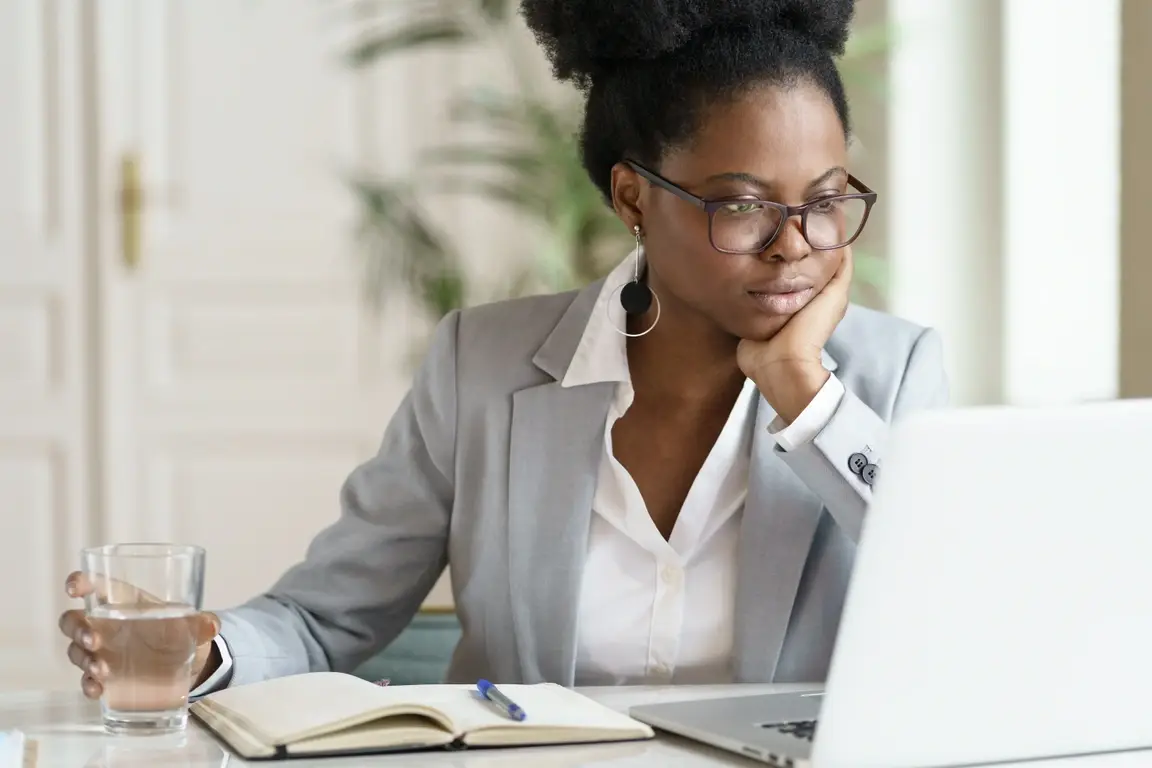 A young black woman working at a desk in front of a computer, looking slightly concerned.