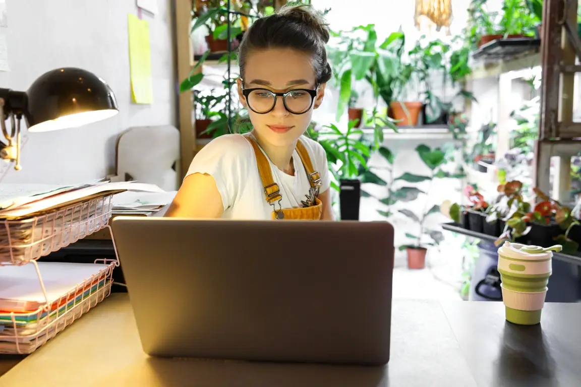 A woman with glasses works on a laptop in a greenhouse with plants behind her and an office setup around her.