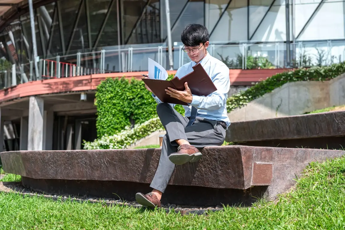 Young man sitting outside an office building looking over documents in a leather folder.