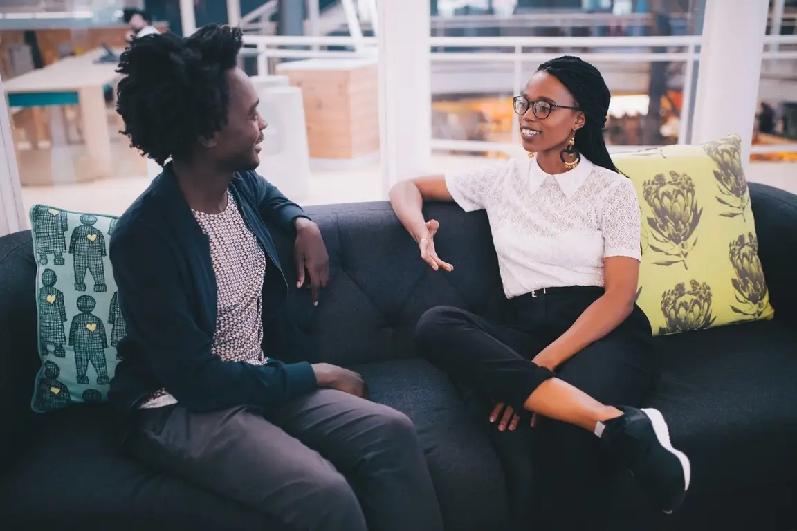 Two young professionals at an office couch having a conversation.