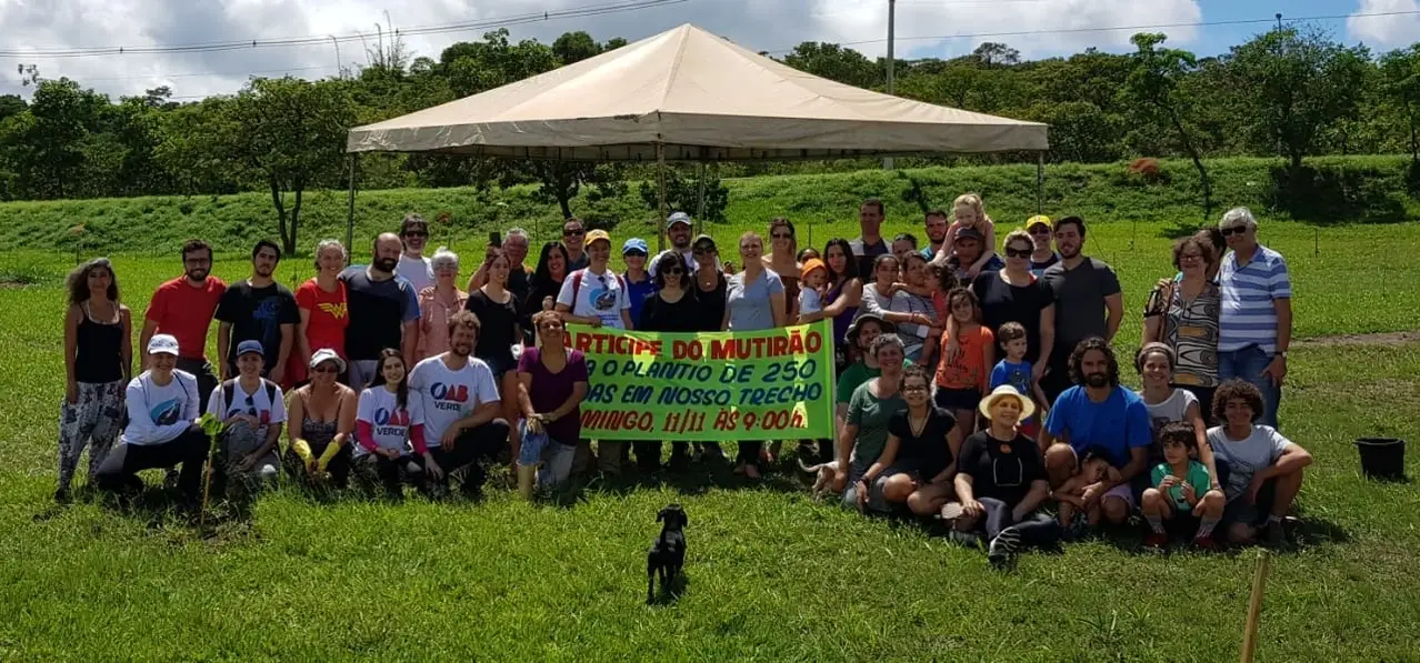 A imagem mostra um grupo de pessoas ao ar livre durante o dia, reunido em uma área gramada com árvores ao fundo, posando para uma foto com uma faixa em português relacionada a um evento.