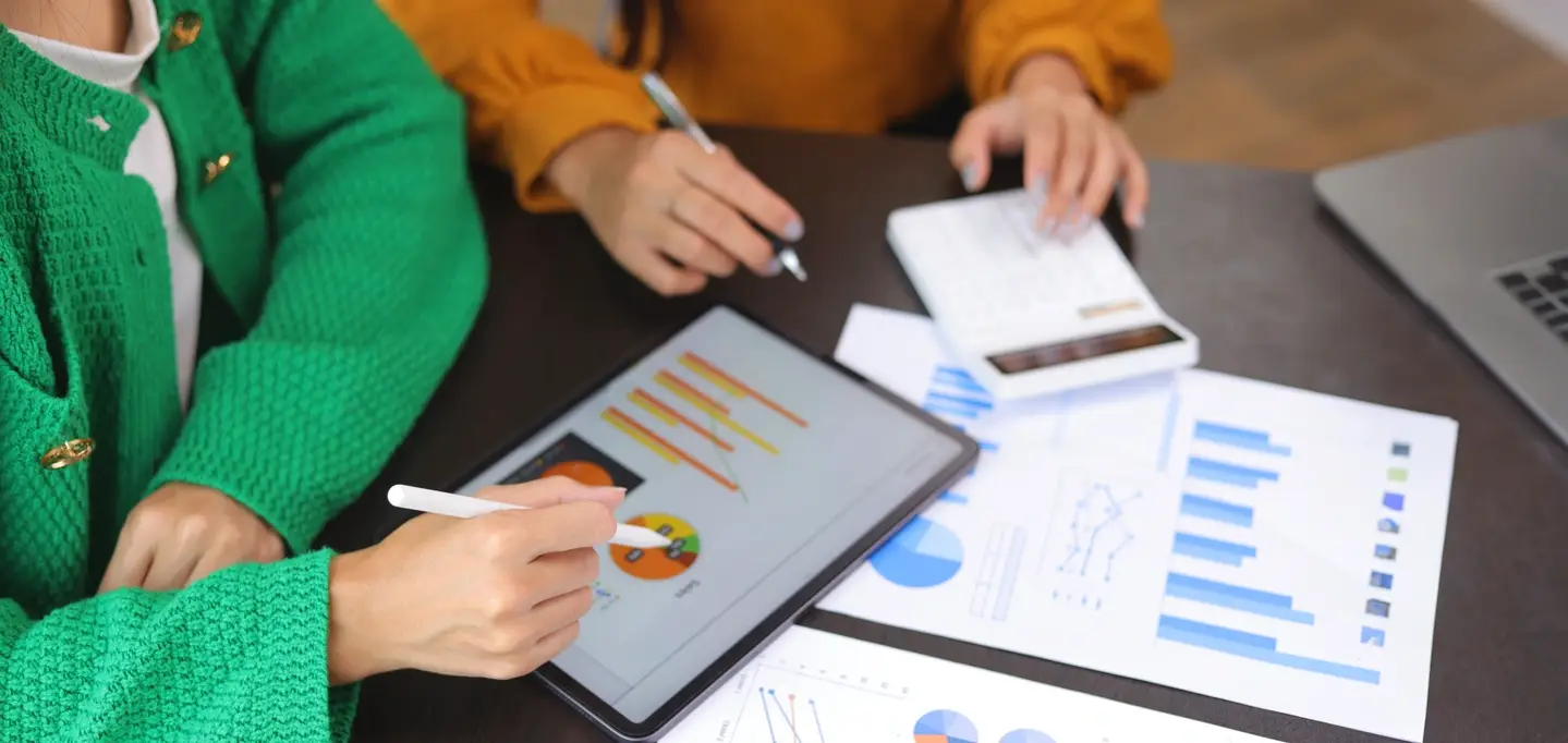 Two co-workers wearing bright sweaters compare research and insights on their tablets and printed out papers.