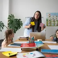 Imagem de uma professora que está segurando cartões coloridos enquanto ensina para um grupo de crianças atentas e sorridentes em uma sala de aula organizada e acolhedora.