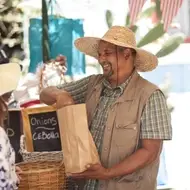 A man wearing a sun hat packages produce at the farmer's market for a customer.