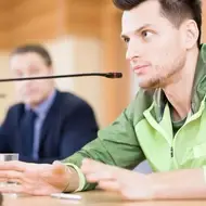 A young white man wearing a green windbreaker sits at a city council meeting to participate in his community.
