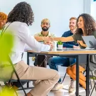 Foto de uma Equipe sentada em uma mesa se cumprimentando.