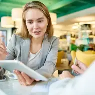 Blonde woman presents work to colleague on an iPad at a coffee shop.