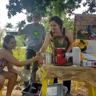 Imagem de 4 pessoas, sendo uma mulher servindo um café à outra, um homem com a camiseta idealista e outra mulher sentada na ponta da mesa.