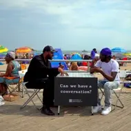 two people sitting at a table on a boardwalk with a sign that reads "can we have a conversation?"