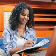 Woman with dark curly hair journals career goals in notebook and while sitting in front of laptop.