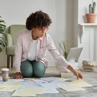 Black woman wearing glasses and button down kneels on rug surrounded by papers.