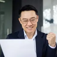 Older Asian man looks at job offer and pumps fist in the air in celebration in office.