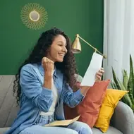 A photograph of a woman with dark curly hair excitedly looking at a piece of paper while sitting on a grey couch.