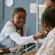 Black woman smiles while treating a small child in an office while wearing scrubs.