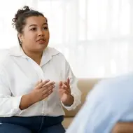 A young woman in white button down speaks to someone sitting across from her while sitting on a couch.