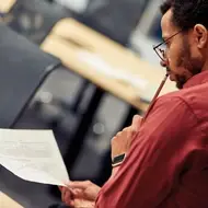 Black man wearing a red shirt and apple watch sits at home looking at grad school student loan paperwork, holding a pencil to his lips.