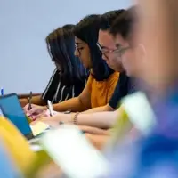 Students in a classroom taking notes with pens and laptops