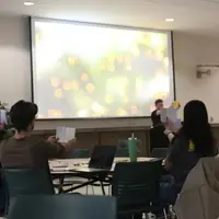 Students in a university setting raise papers to critique a presentation with soft bokeh lights projected in the background.