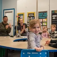 A group of adults and children sit at a round table. One of the adults is signing. A young girl turns to smile at the camera.