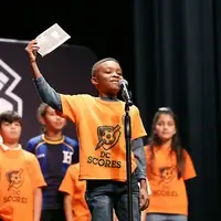 young Black boy speaking into a mic with his teammates on stage behind him