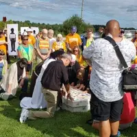 Clergy kneel as they pray over a mock casket with pictures of undocumented people they know are attached to the outside. They are surrounded by others in orange LA RED shirts with media present.