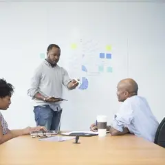 A group of three professionals are engaged in a collaborative meeting. A man standing at the whiteboard gestures while holding a tablet, leading the discussion. Another man, seated at the table, listens attentively with a coffee cup and pen in front of him. A woman sits across from him, focused on her laptop, contributing to the conversation. The whiteboard behind them displays charts, sticky notes, and brainstorming ideas, suggesting a strategic planning or business development session.