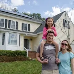 Family of three stands in front of a home with solar. The young girl sits on her dad's shoulders.