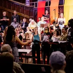 A group of ten teens and young adults are in the foreground, performing their songs and spoken word pieces. The main choir is seated in the background.