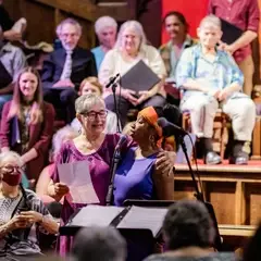 Two choir members, one wearing a pink shirt and glasses, the other with a purple tank top and orange headband, stand at the microphone. The full choir is seated on the stage in the background.