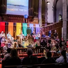 The full choir of 50 or more members are seated on the stage. A guest performer dances with an inflatable globe on a second stage, above the choir. A row of six banners (red, orange, yellow, green, blue, purple) hang from the second stage.