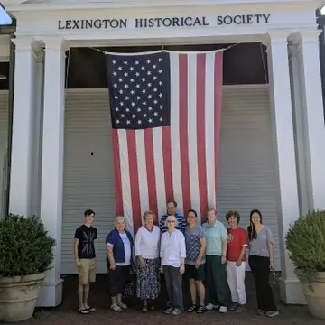 Group of people standing in front of an American flag suspended from the roof of a large, white building labelled "Lexington Historical Society"
