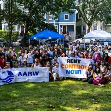 photo of a group of people, mostly Asian American, standing and smiling with banners that read "AARW" and "Rent Control Now"