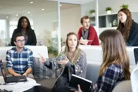 Foto de 6 pessoas em uma sala de discussão, com fundo branco, cheio de plantas e com porta de vidro. Três pessoas sentadas em volta de uma mesa cheia de papéis, uma caneta e um computador. Três pessoas em pé, todos com virados para mesa, parecem estar participando da discussão.