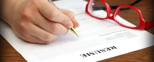 Someone writing with a pen on a resume. There are red glasses on the desk as well.