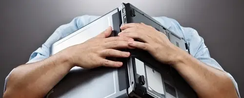 A distressed man holds an open laptop over his head.