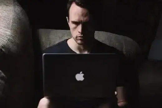 Um homem sentado em um sofá olhando para seu laptop.