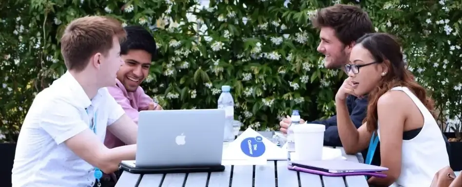 Imagem de 4 jovens reunidos ao redor de uma mesa de madeira, com um notebook, cadernos e garrafas de água.