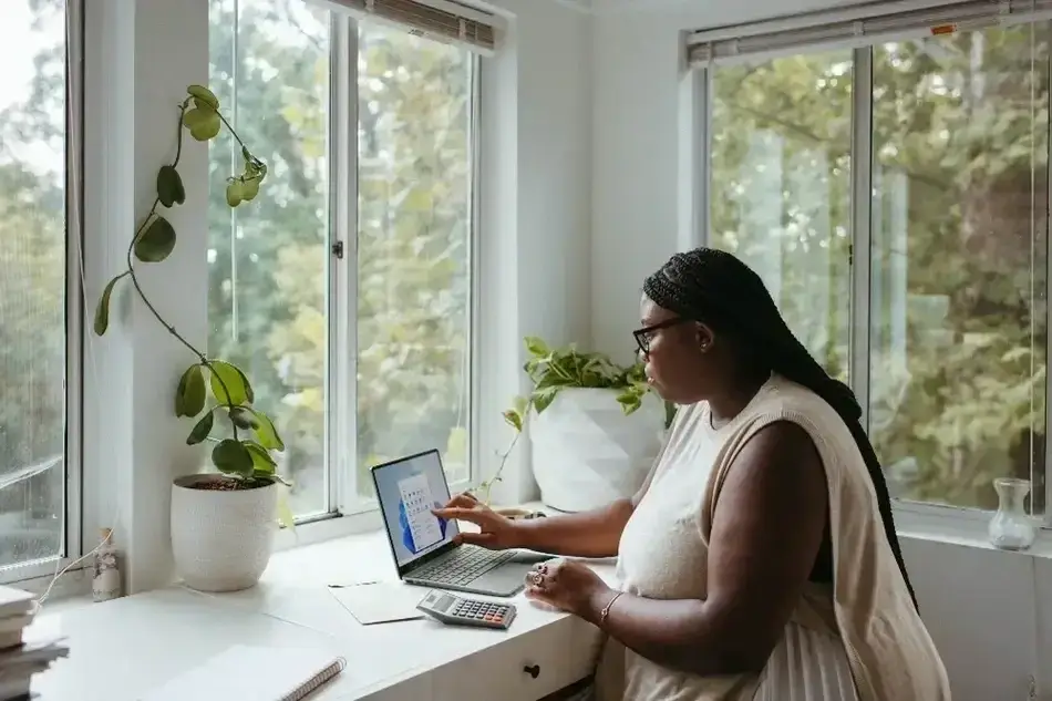 Imagem de uma mulher afrodescendente de branco e faz contraste com a sala que é toda branca. Ela está sentada na frente de um notebook com uma calculadora, alguns papéis. Está em uma mesa próximo da janela, é uma sala bem iluminada e cheia de plantas que podemos ver através da janela e dentro da sala em vasos.