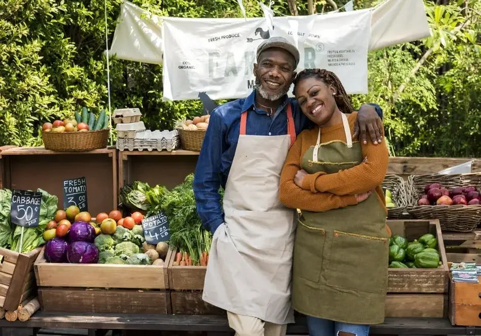 Um homem e uma mulher negros estão juntos, sorrindo em frente a uma barraca de alimentos frescos para destacar os benefícios de se voluntariar em uma feira agrícola.