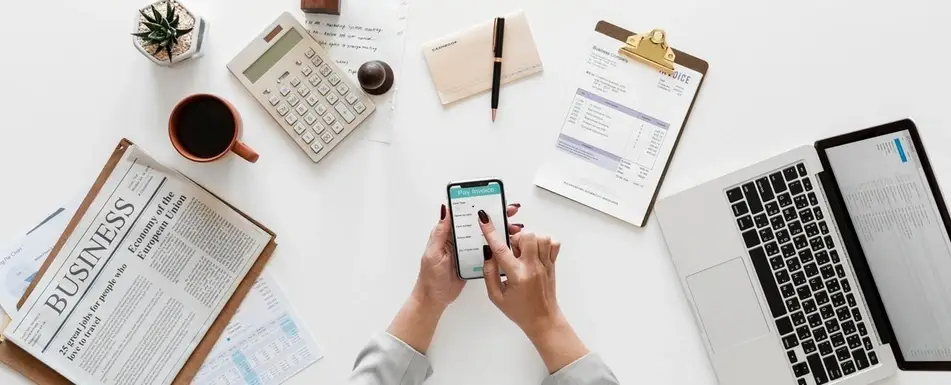 A person using their smartphone. On their desk is their laptop, a calculator, a newspaper, a cup of coffee, a pen and a clipboard