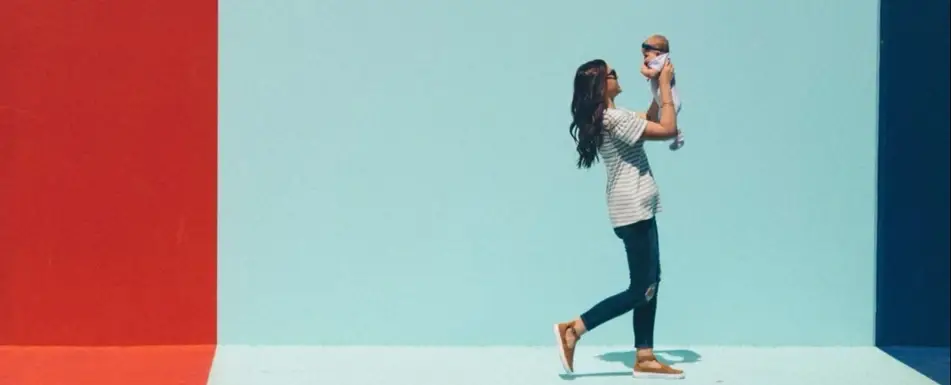 A woman standing in front of a colorful wall holds up a baby.