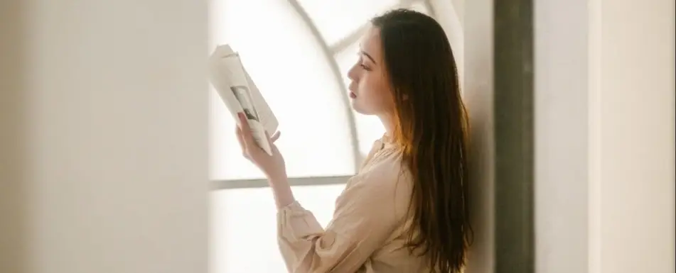 A woman reads a pamphlet by a window.