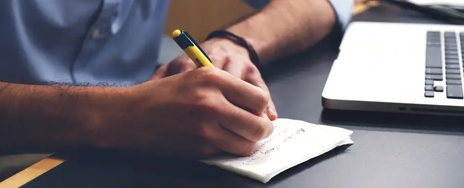 Someone writing on a pad of paper next to a laptop.