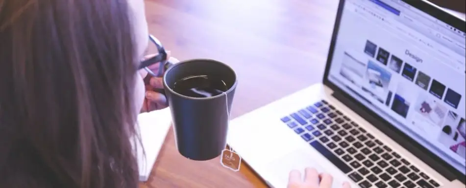 A woman works at a laptop with a mug of tea in hand.