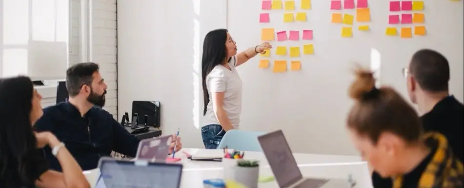 A group of people ina conference room. One of them is pointing at a whiteboard. The board has a lot of post its on it.