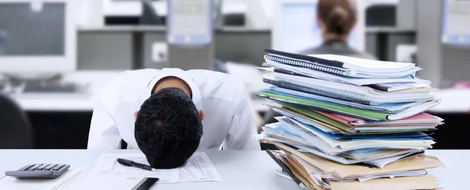 A person has their head on their work desk. There's a pile of papers and books next to them.