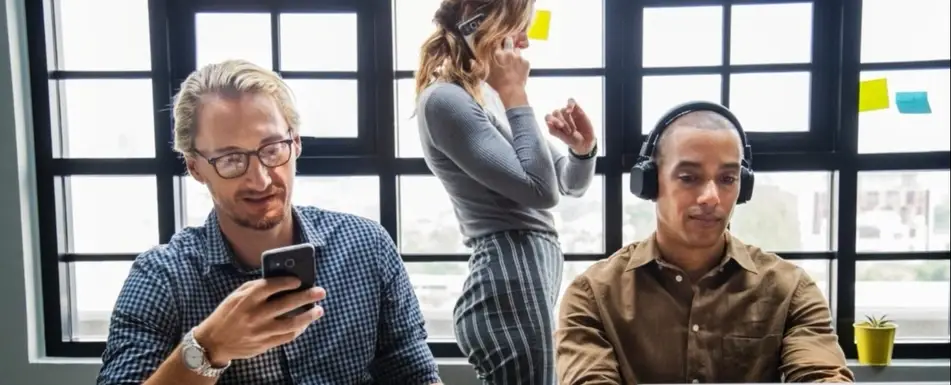 Three people in the office. One is wearing headphones, the other is on a phone call and the other is looking at their phone.