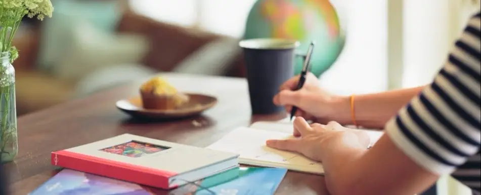 A person writing in a notebook. There are other books and a muffin on the table as well.