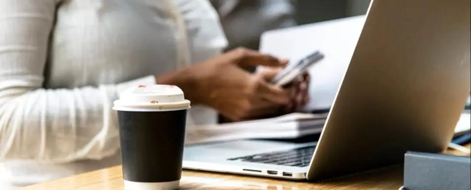 A close up of a cup of coffee next to someone working at a laptop.